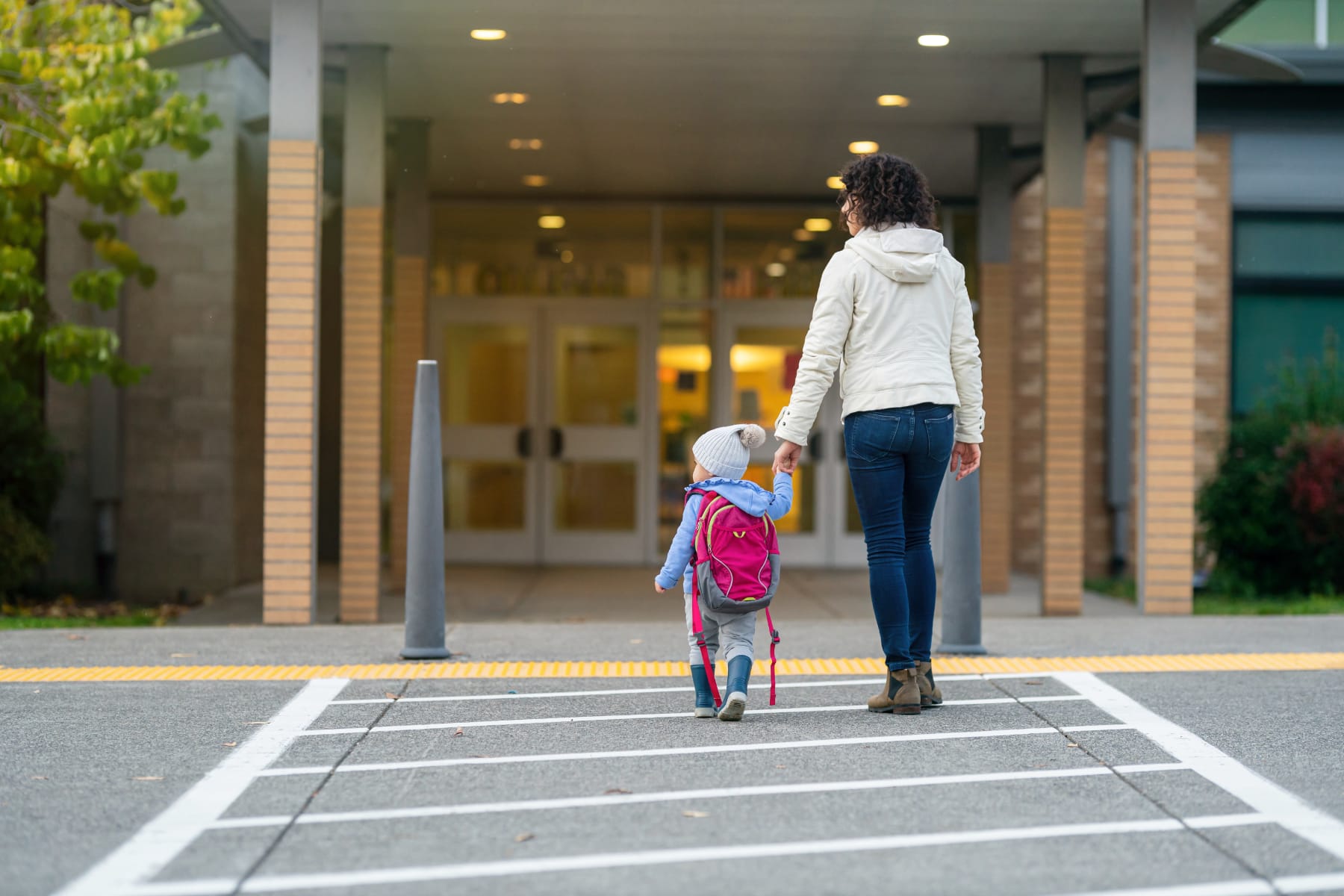 a woman holds the hand of a toddler who has a backpack on; they are seen from behind walking into a building