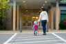 a woman holds the hand of a toddler who has a backpack on; they are seen from behind walking into a building