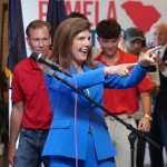 Pamela Evette, in a blue suit, points while standing in front of a crowd at a rally
