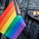 A close up of part of a denim jacket with a Pride pin on the pocket and a Pride flag
