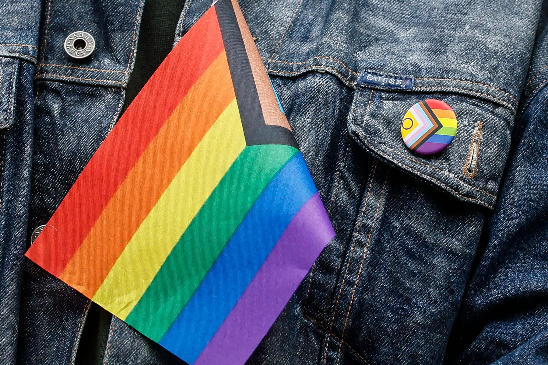 A close up of part of a denim jacket with a Pride pin on the pocket and a Pride flag