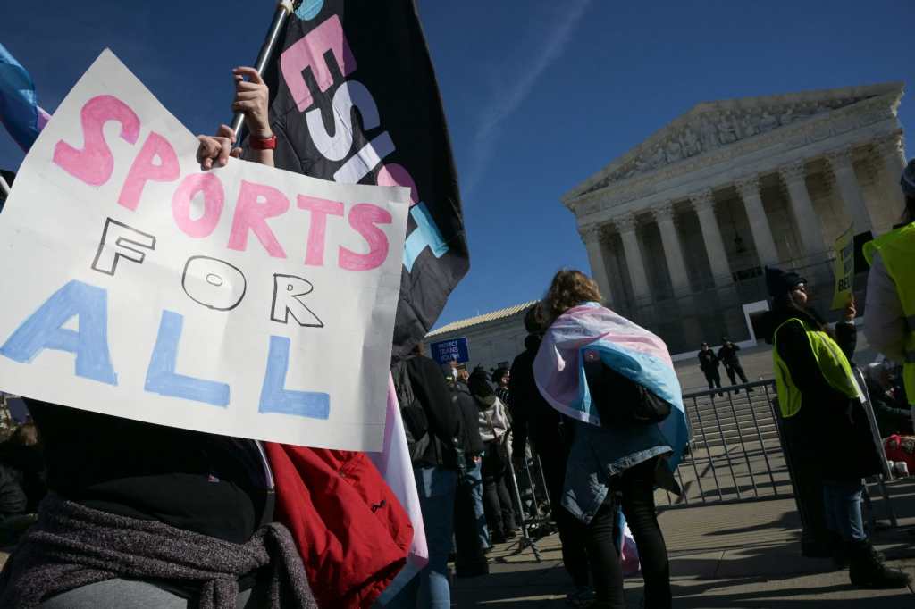 A person holes a sign that says "sports for all" in pink, white and blue, another holds a flag with "resist" in pink, white and blue against a black background, and another is wrapped in a trans Pride flag in front of the Supreme Court