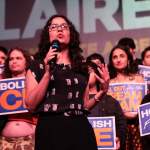 Claire Valdez speaks with a crowd behind her holding signs that says Housing for All and Abolish ICE