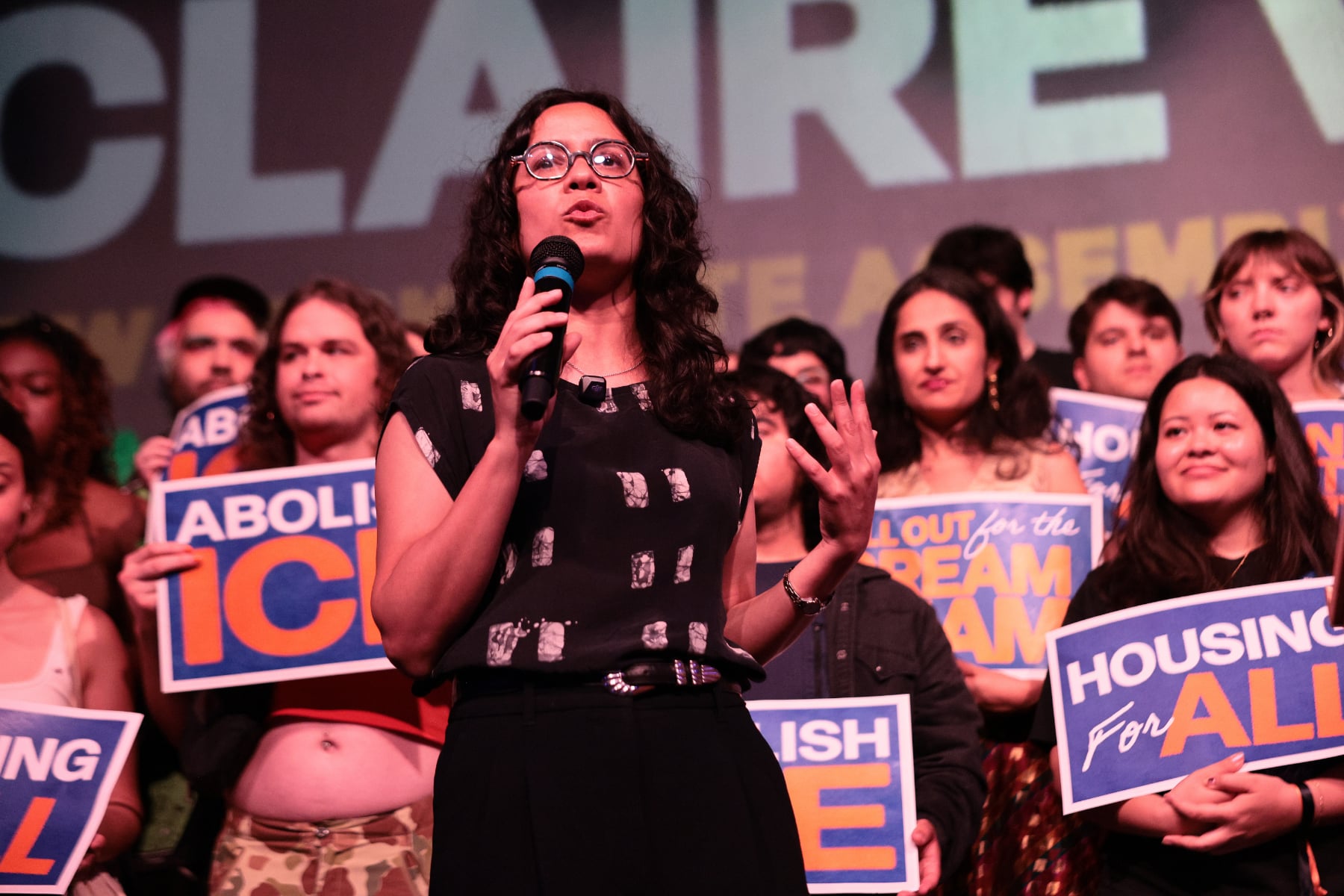 Claire Valdez speaks with a crowd behind her holding signs that says Housing for All and Abolish ICE