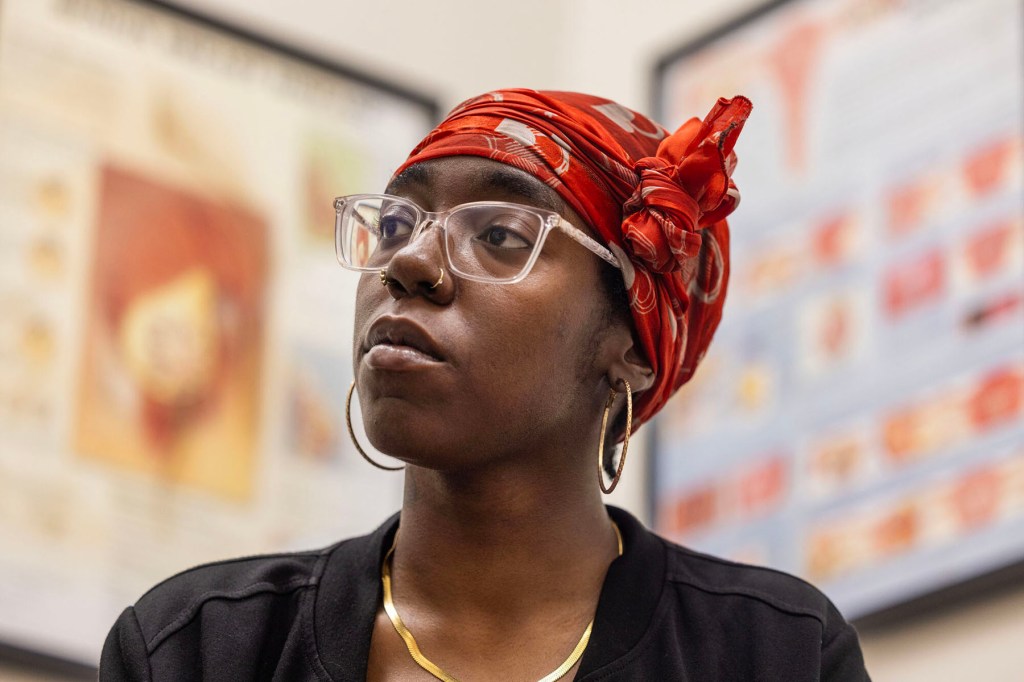 A woman wearing clear glasses and a red patterned headwrap looks on in a medical exam room.