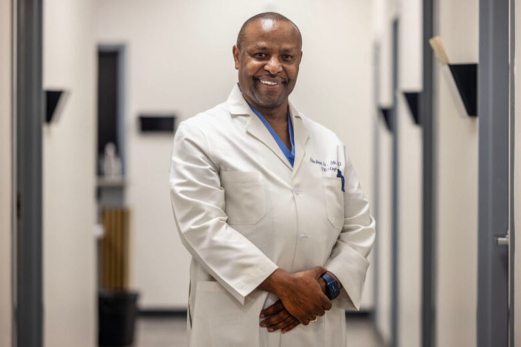 A smiling doctor in a white coat stands with his hands clasped in a clinic hallway.
