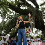 Annie Andrews speaks into a microphone and raises one hand while addressing a crowd gathered beneath a large oak tree with American flags and protest signs.