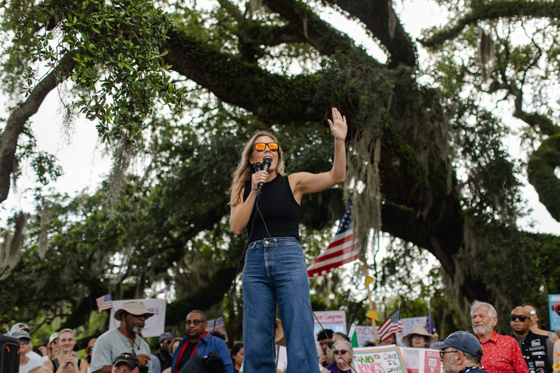 Annie Andrews speaks into a microphone and raises one hand while addressing a crowd gathered beneath a large oak tree with American flags and protest signs.