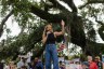 Annie Andrews speaks into a microphone and raises one hand while addressing a crowd gathered beneath a large oak tree with American flags and protest signs.