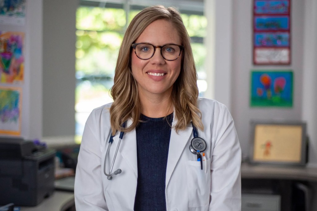 A smiling Annie Andrews wearing glasses, a white medical coat and a stethoscope while posing in an office decorated with colorful children's artwork.