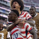 Folarin Balogun celebrates on the field as teammates crowd around him, shouting and embracing after a goal.