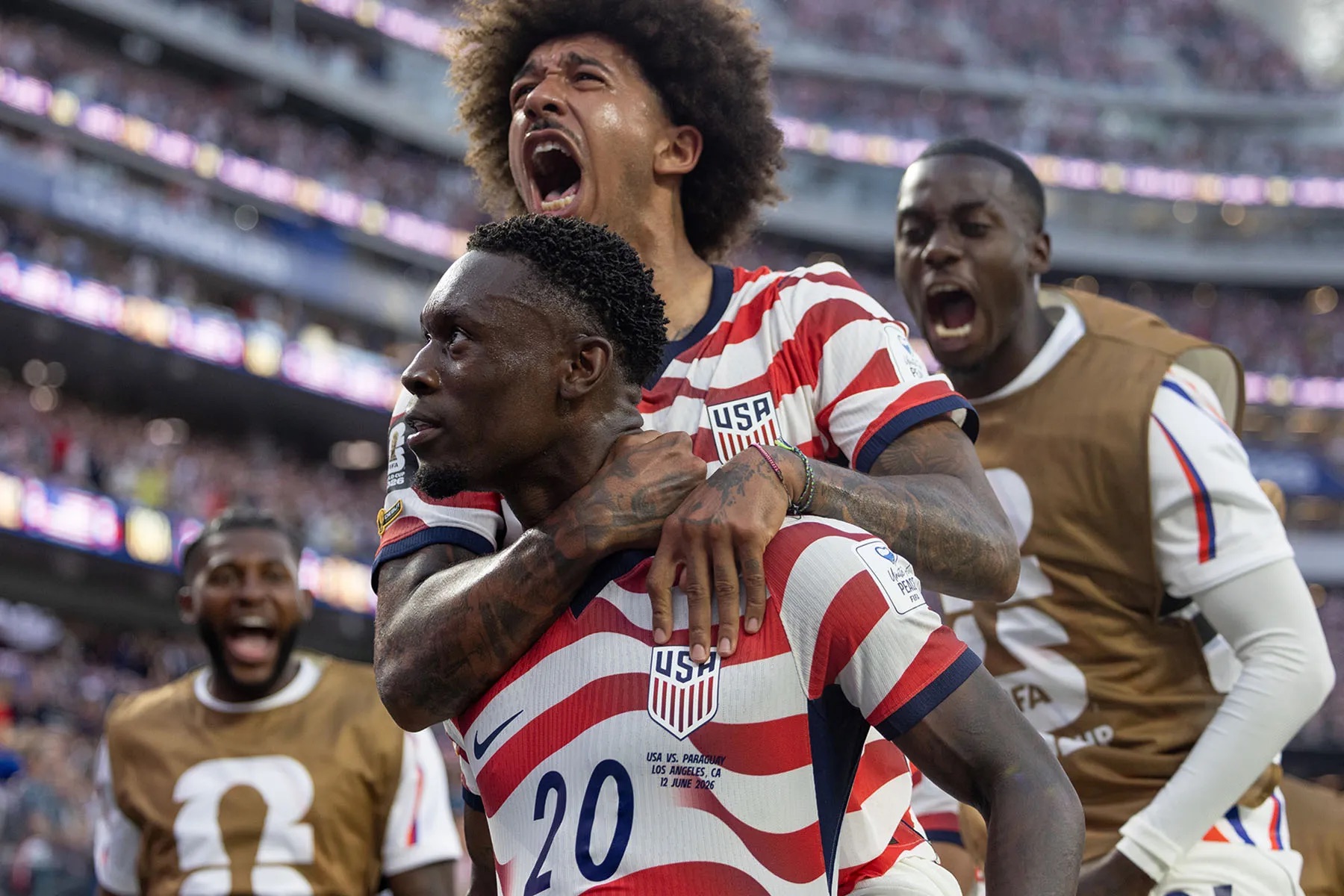 Folarin Balogun celebrates on the field as teammates crowd around him, shouting and embracing after a goal.