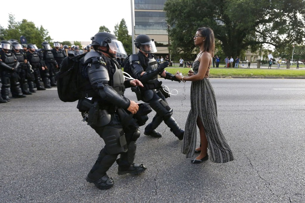 A woman in a dress stands calmly in the street facing a line of police officers in riot gear. Two officers move toward her with their arms out as more officers stand behind them.