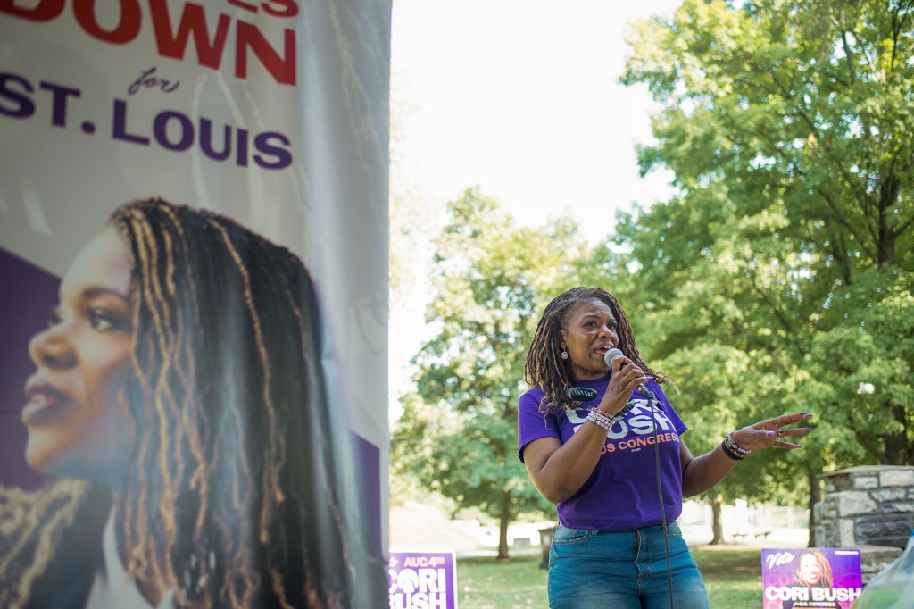 Cori Bush speaks into a microphone at an outdoor campaign event beside a large banner featuring her portrait.