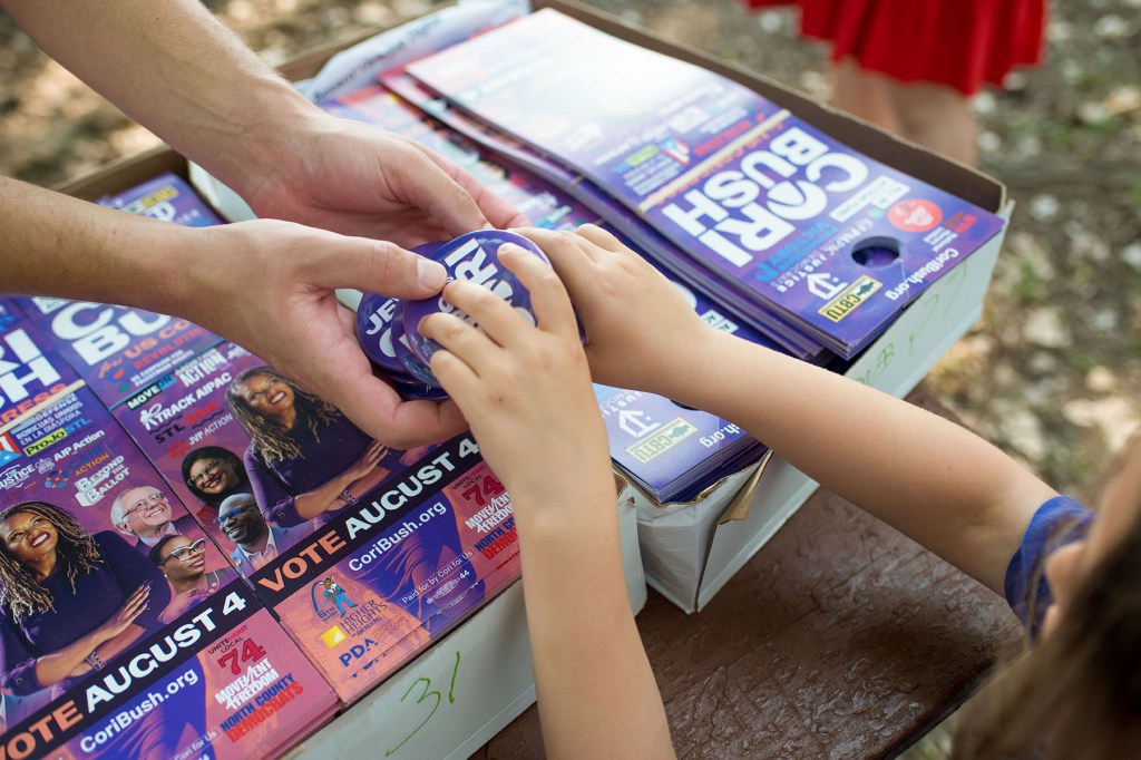 A child reaches for a Cori Bush campaign button above a table covered with flyers and other campaign materials.