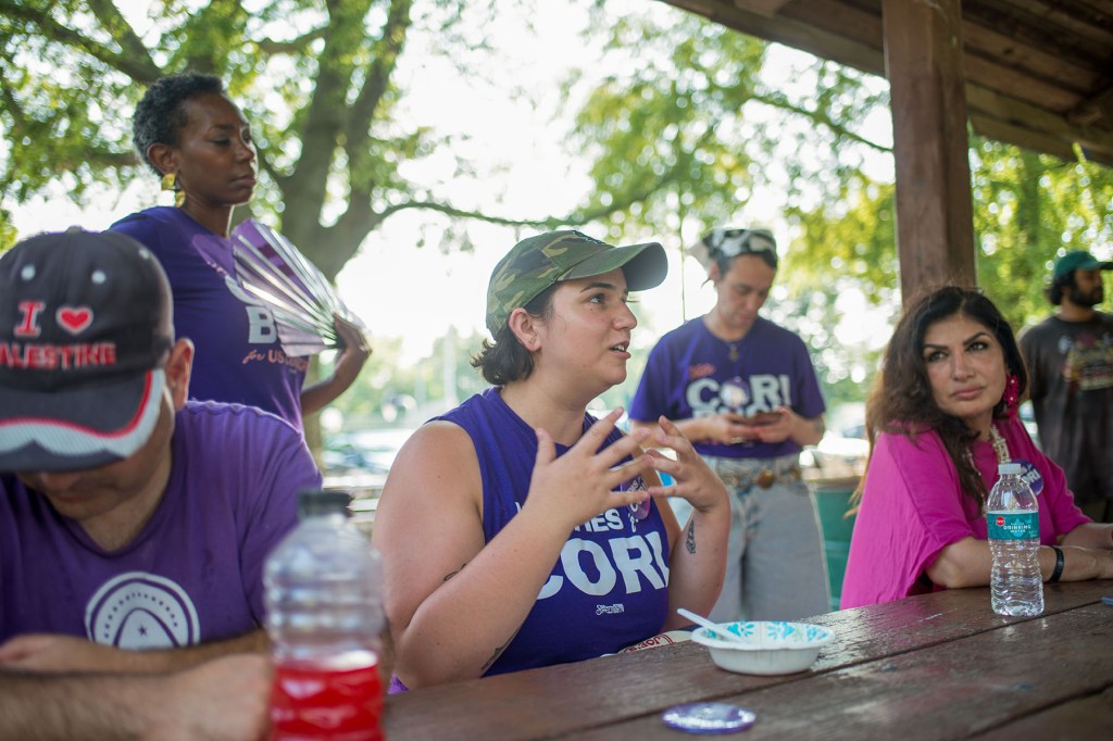 Cori Bush campaign volunteers gather around a picnic table as one person speaks, with supporters wearing purple campaign shirts nearby.