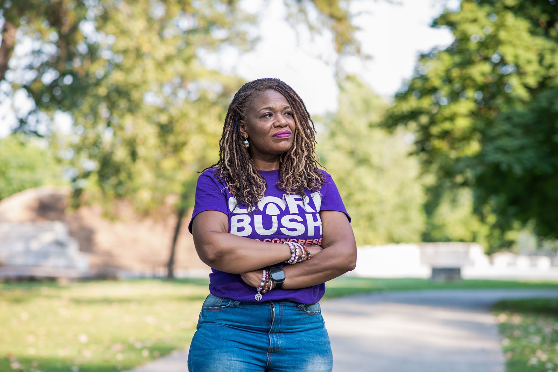 Cori Bush stands with her arms crossed in a park, wearing a purple campaign T-shirt and looking off to the side.
