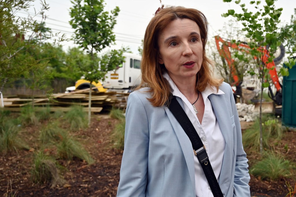 A woman in a light blue blazer speaks outdoors near construction equipment.