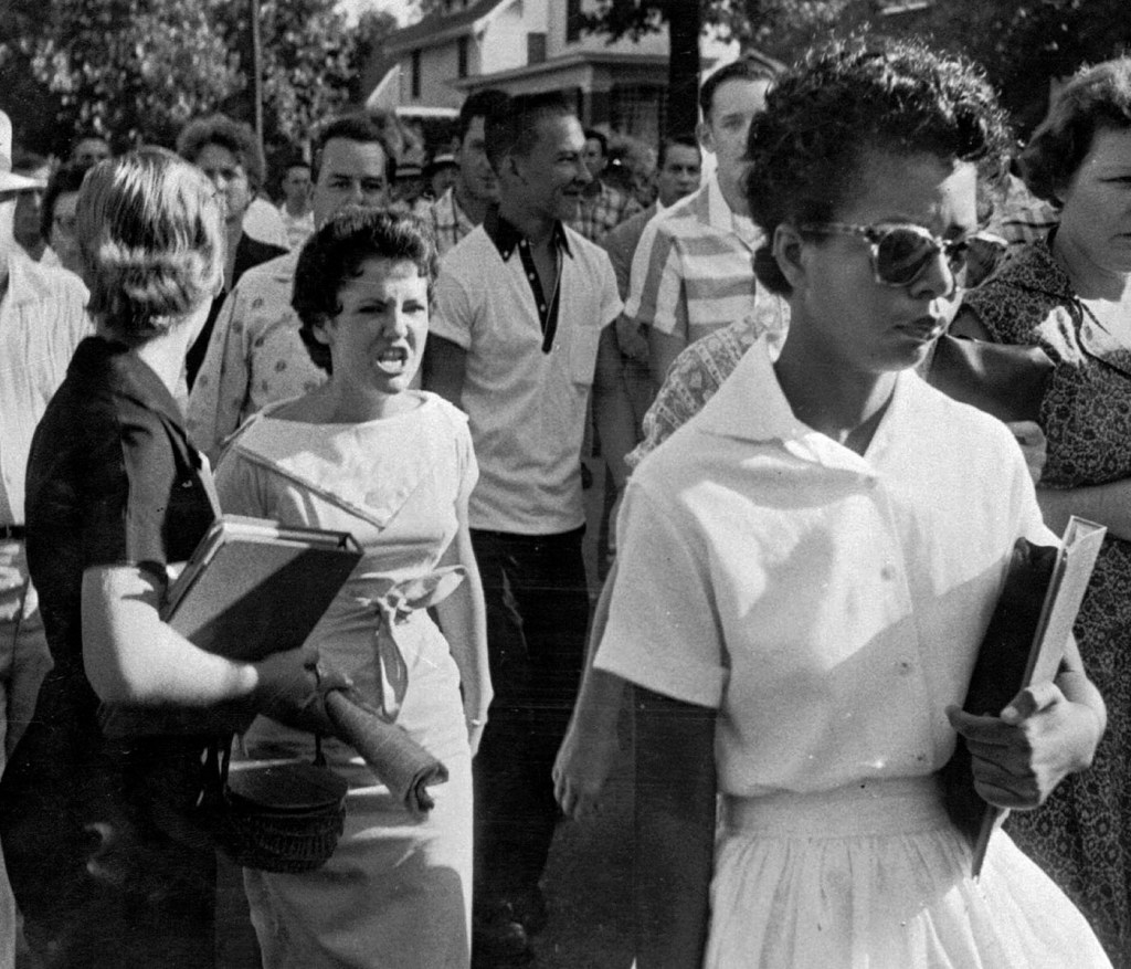 Black and white photo of Elizabeth Eckford, a Black teenage girl wearing sunglasses and carrying books, walking past a crowd outside Little Rock Central High School. Behind her, Hazel Bryan, a white student, shouts at her as others look on.