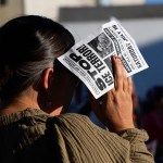 A demonstrator holds a protest flyer over their face to block the sun. The flyer reads “Stop ICE Terror!” and calls for justice for Lorenzo and Joan and the jailing of ICE agents.