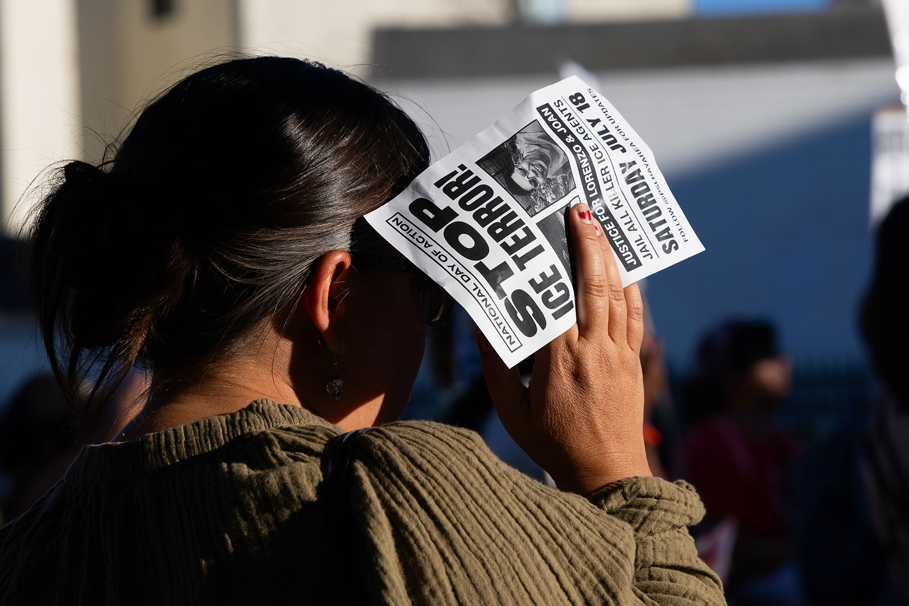 A demonstrator holds a protest flyer over their face to block the sun. The flyer reads “Stop ICE Terror!” and calls for justice for Lorenzo and Joan and the jailing of ICE agents.
