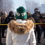 A person in a white parka and green knit hat faces a line of heavily equipped federal law enforcement officers wearing gas masks behind yellow police tape on a smoky Minneapolis street.