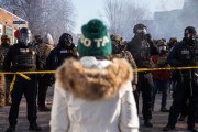 A person in a white parka and green knit hat faces a line of heavily equipped federal law enforcement officers wearing gas masks behind yellow police tape on a smoky Minneapolis street.