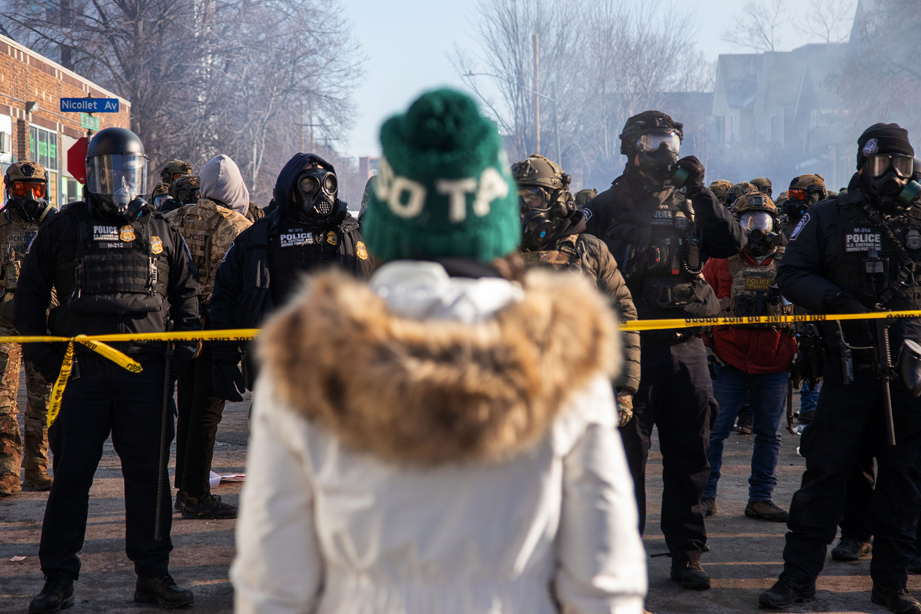 A person in a white parka and green knit hat faces a line of heavily equipped federal law enforcement officers wearing gas masks behind yellow police tape on a smoky Minneapolis street.