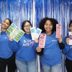 Four young people wearing matching blue shirts pose in front of a metallic blue fringe backdrop while holding colorful pamphlets about consent, sexual health education and LGBTQ+ inclusion.