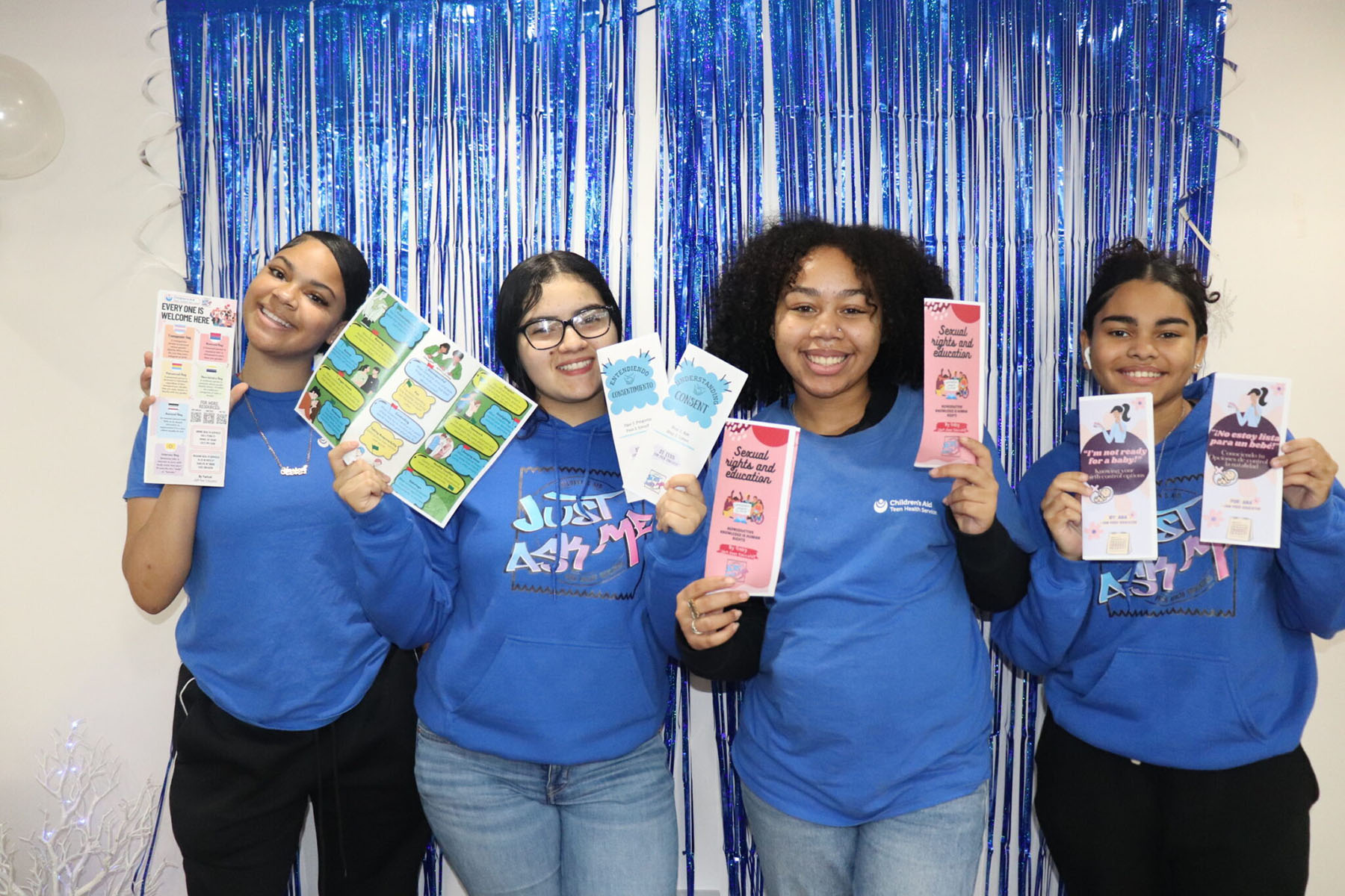 Four young people wearing matching blue shirts pose in front of a metallic blue fringe backdrop while holding colorful pamphlets about consent, sexual health education and LGBTQ+ inclusion.