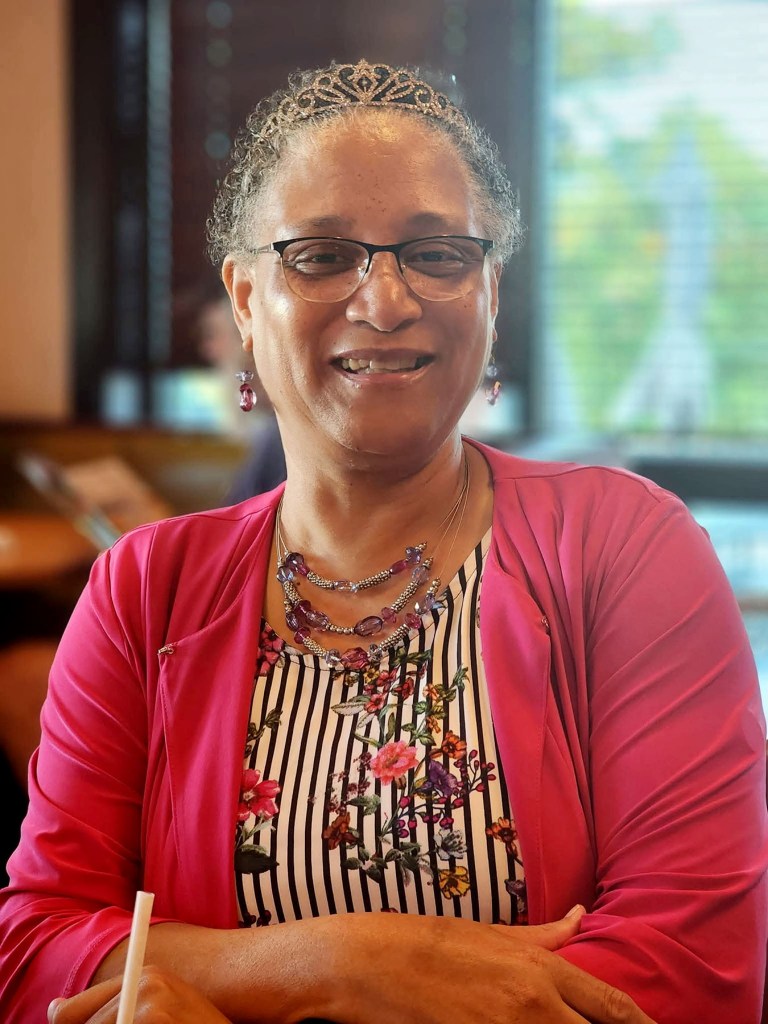 Julie Chenault smiles while seated indoors, wearing glasses, a tiara, a bright pink cardigan and layered purple necklaces.