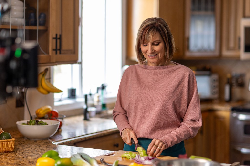 Kerry Dennis smiles as she chops vegetables on a cutting board in a kitchen.