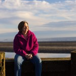 LauraLee Denler, wearing a bright pink fleece and jeans, sits outdoors near a beach and looks to the side.