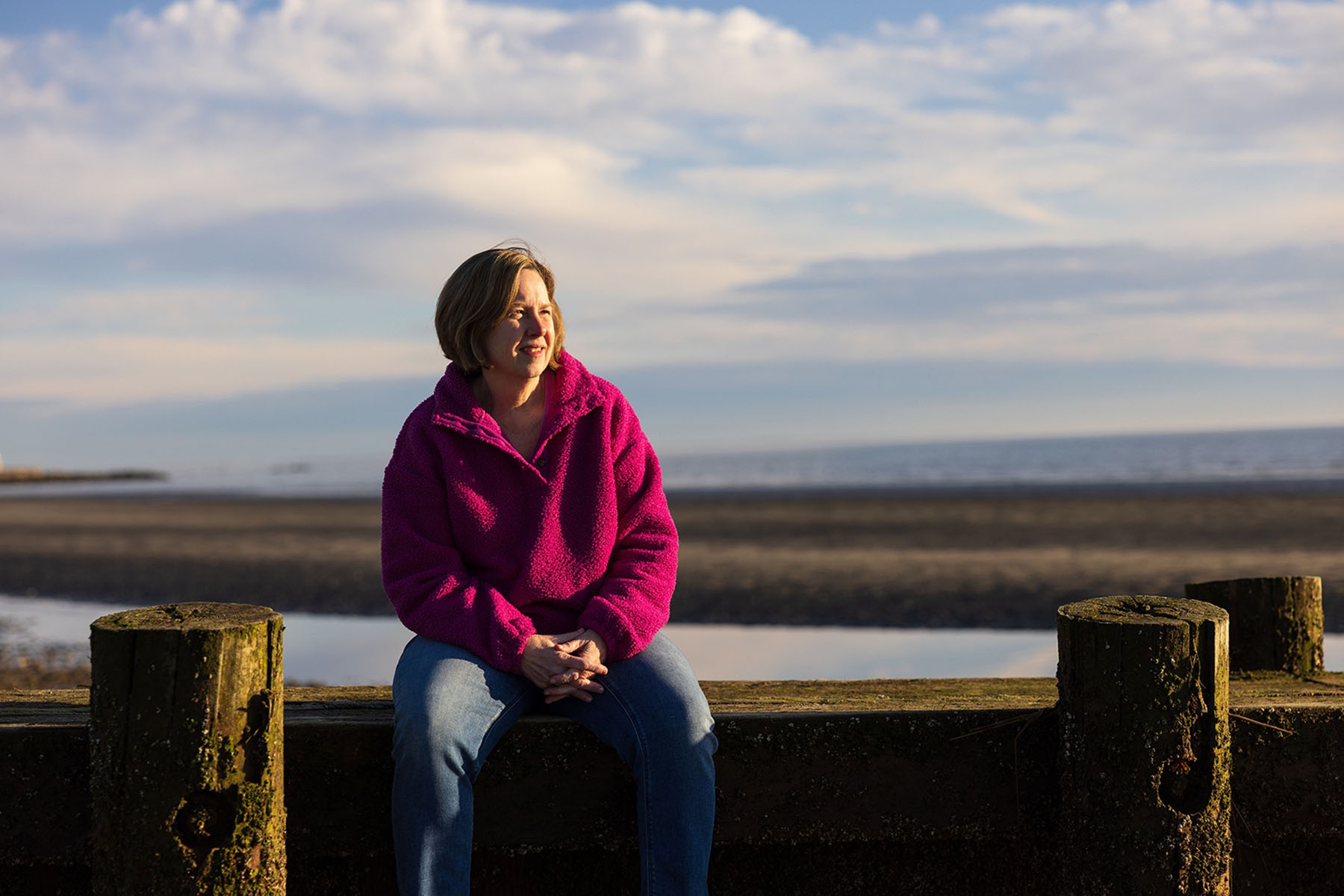 LauraLee Denler, wearing a bright pink fleece and jeans, sits outdoors near a beach and looks to the side.