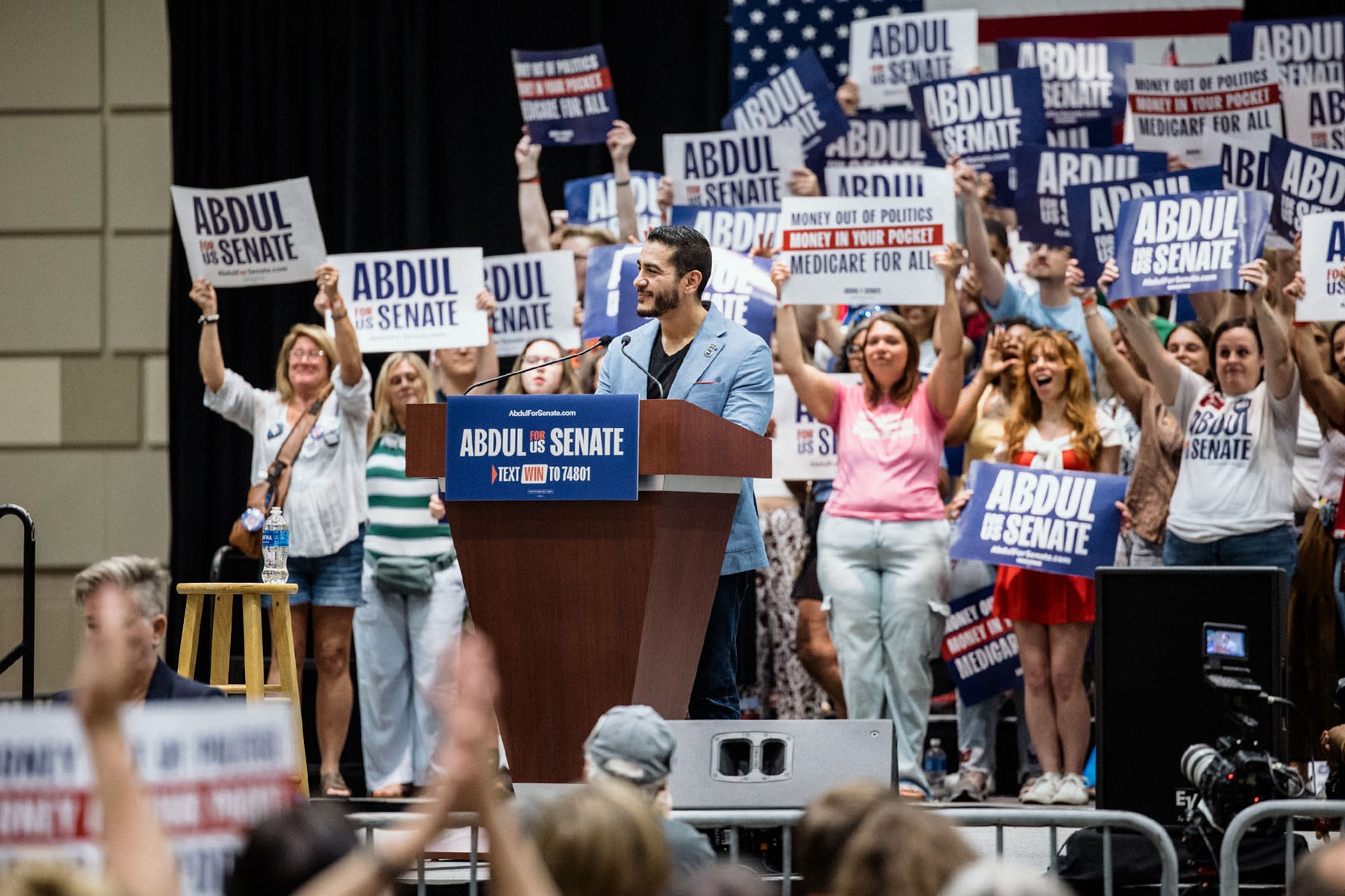 Senate candidate Abdul El-Sayed smiles at a podium as supporters behind him raise campaign signs.