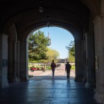 Two students walk through a shadowed archway onto the Texas Tech University campus, with trees, flags and campus buildings visible in the distance.