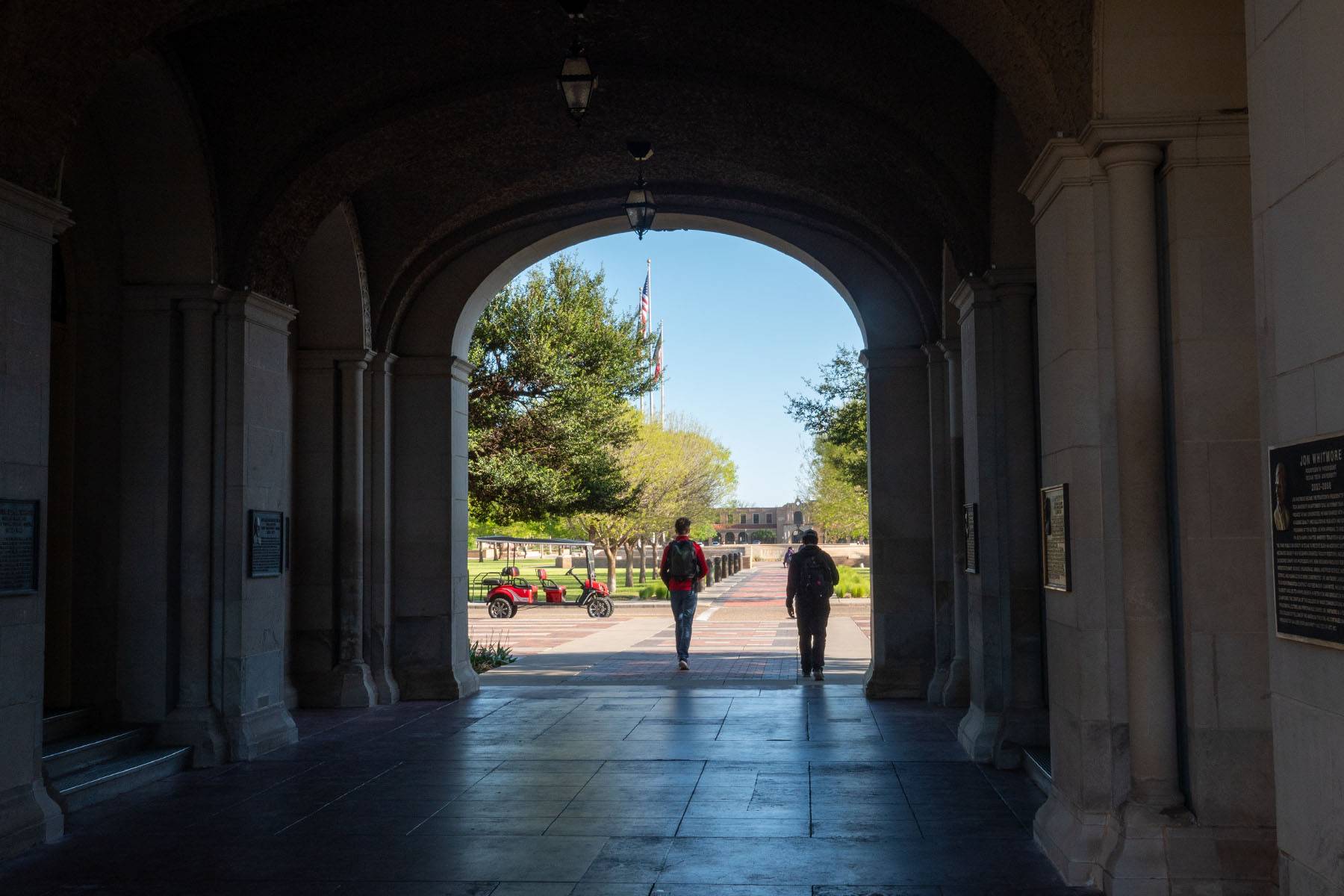 Two students walk through a shadowed archway onto the Texas Tech University campus, with trees, flags and campus buildings visible in the distance.