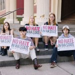 Five young people sit on the steps of a building, each holding signs that read, “No hate in WA state. Vote No on I-001 & I-638.”