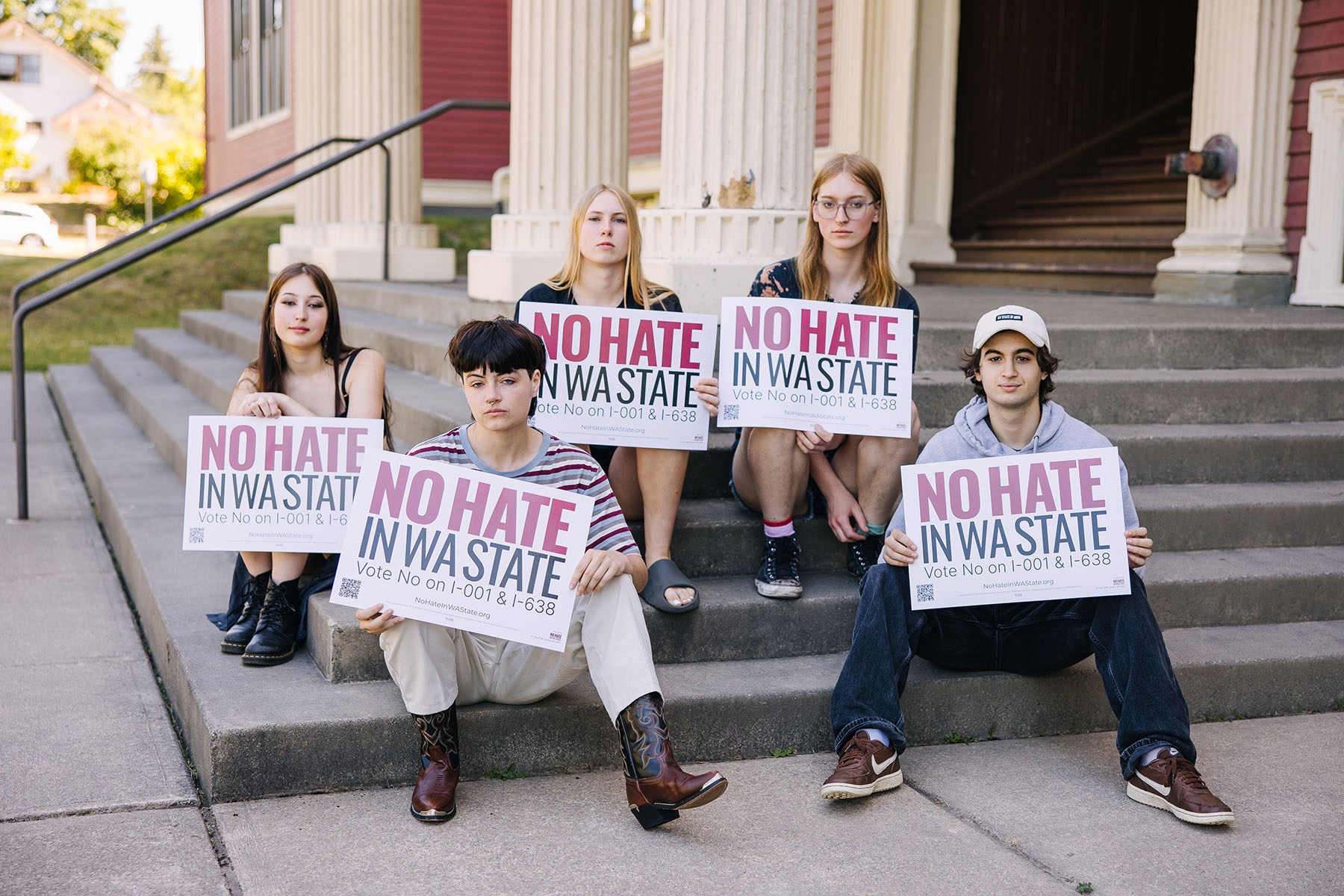 Five young people sit on the steps of a building, each holding signs that read, “No hate in WA state. Vote No on I-001 & I-638.”