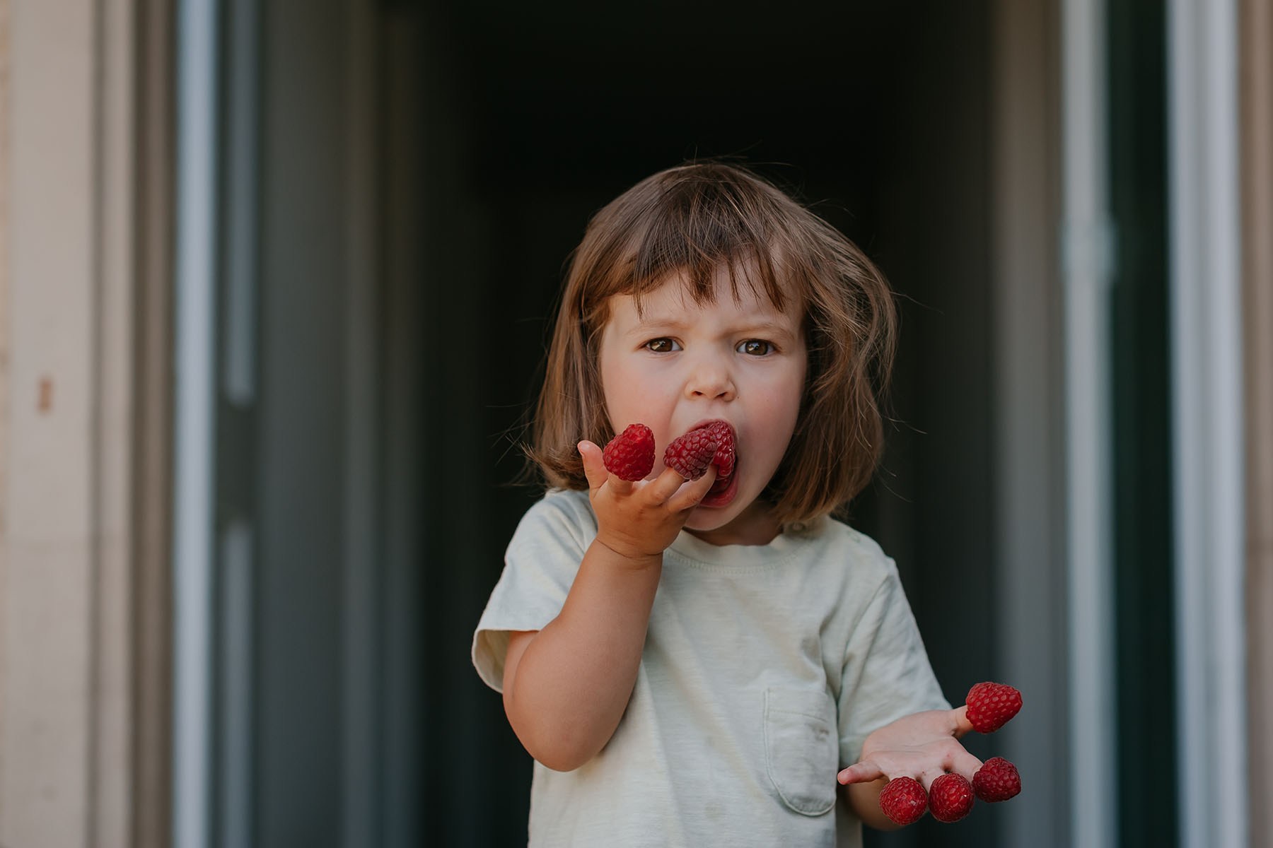 A young child stands in a doorway, holding several raspberries and eating two from one hand.