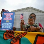 A woman holds her baby and an America flag in front of the Supreme Court alongside someone holding a sign saying American Born Children are American Children.