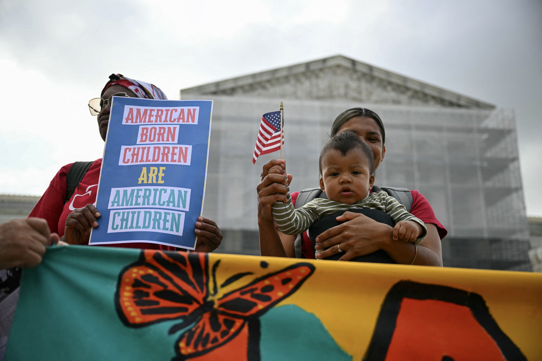 A woman holds her baby and an America flag in front of the Supreme Court alongside someone holding a sign saying American Born Children are American Children.