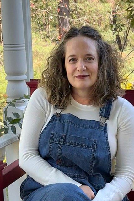 A woman wearing overalls and a white shirt smiles at the camera, seated outside on a bench.