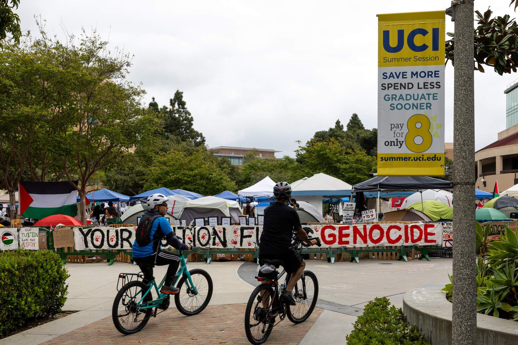 Two cyclists ride by a protest encampment with signage showing support for Palestine and a UCI sign on a light pole nearby.