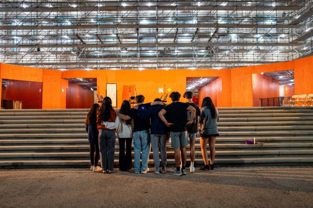 A group of students huddle together holding each other in front of a campus building.