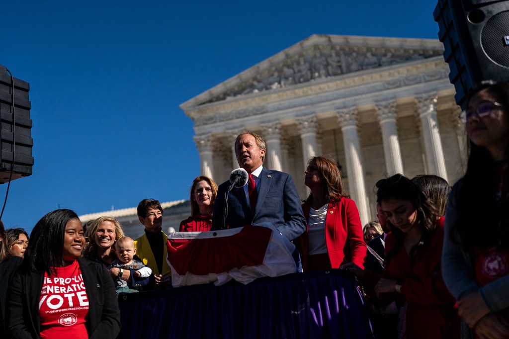 Ken Paxton speaks at a podium outside the Supreme Court surrounded by anti-abortion advocates.
