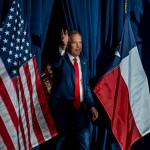 Ken Paxton, wearing a navy suit and red tie, waves while walking onstage between an American and a Texas flags.