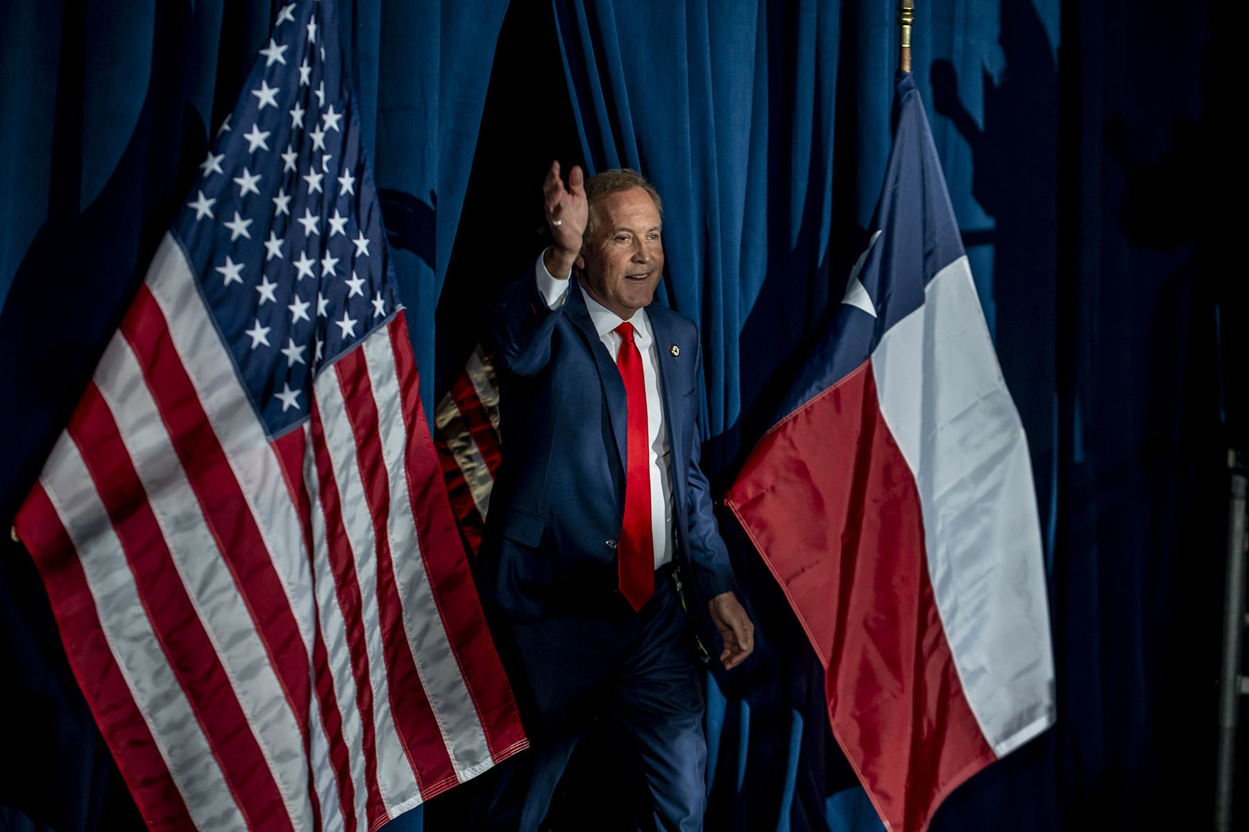 Ken Paxton, wearing a navy suit and red tie, waves while walking onstage between an American and a Texas flags.