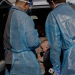 Two health care workers wearing blue protective gowns and face shields lean into an open vehicle door to examine a seated woman holding a baby.