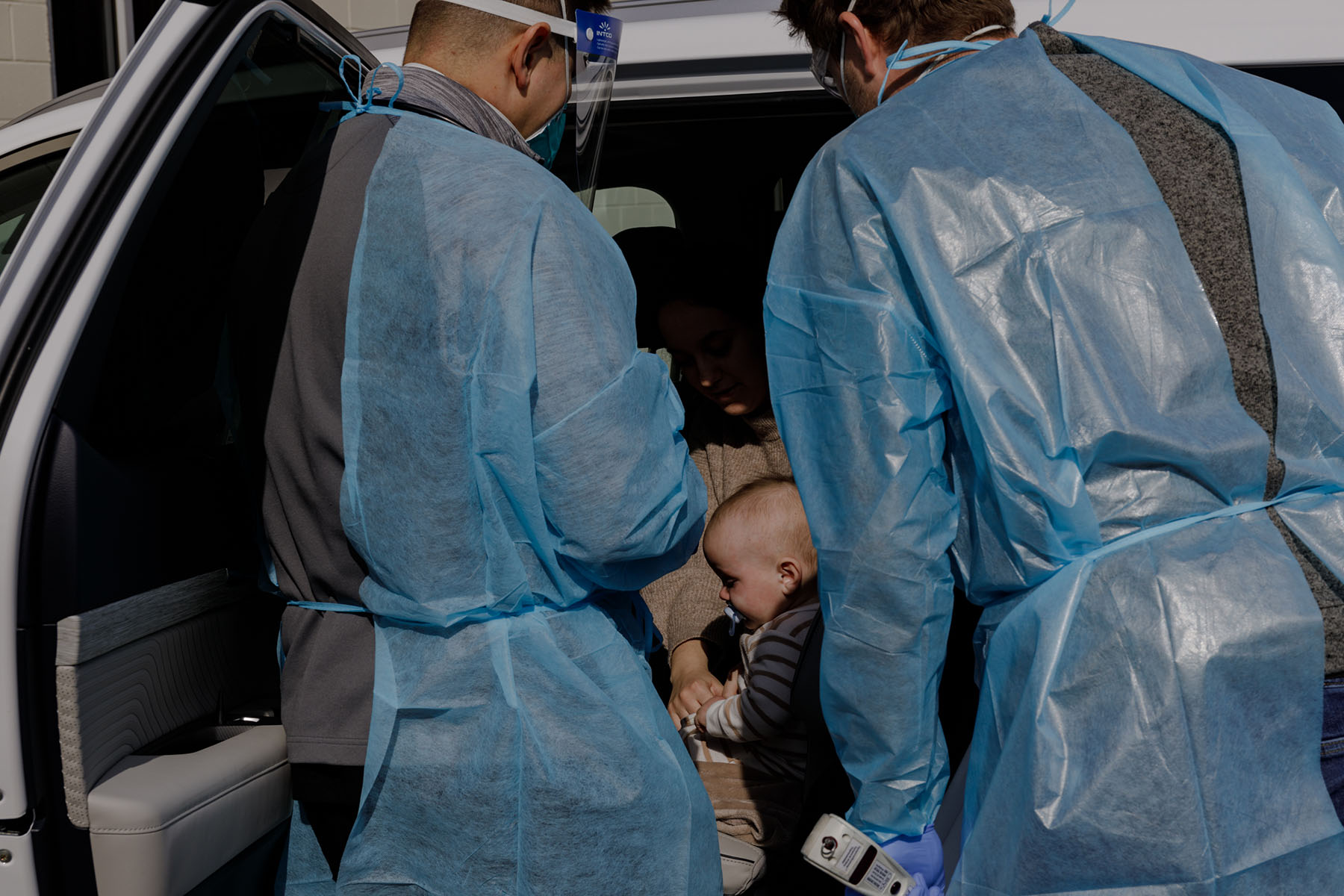 Two health care workers wearing blue protective gowns and face shields lean into an open vehicle door to examine a seated woman holding a baby.
